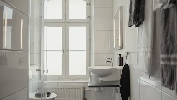 A bright, modern bathroom with white tiles, a large window, a sink, and fresh towels.