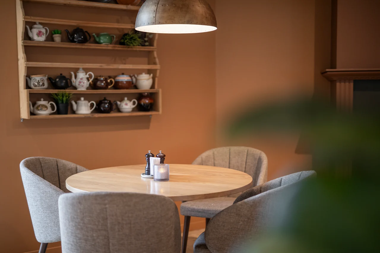 A round wooden table with salt and pepper shakers and a candle stands surrounded by gray chairs. Shelves with various teapots hang on a brown wall under a metal pendant light.