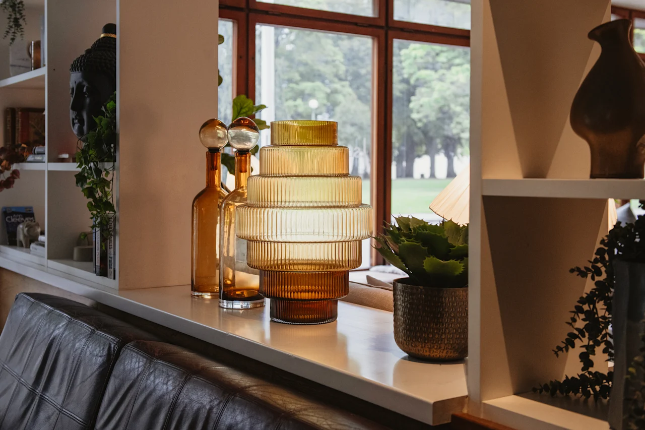 A cozy indoor scene featuring a tiered amber glass lamp, decorative bottles, and potted plants by a large window overlooking lush greenery.