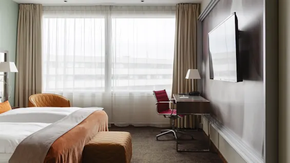 Ett modernt hotellrum med en säng, ett skrivbord, en röd stol och ett stort fönster med gardiner. En TV är monterad på väggen.
