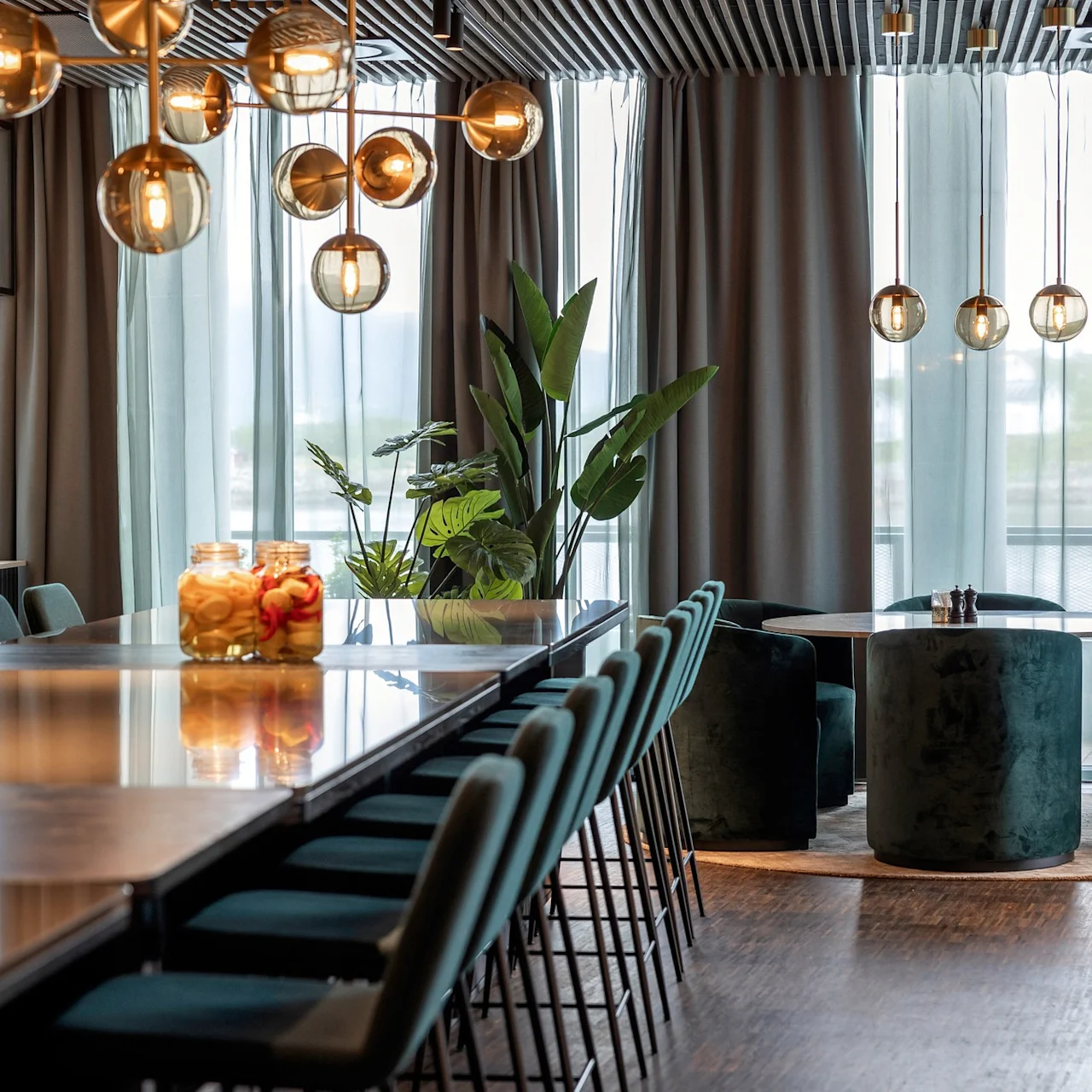 Elegant spiseområde med et langt bord og grønne stole under dekorative pendellamper. Store vinduer og indendørs planter giver en moderne, luftig atmosfære.