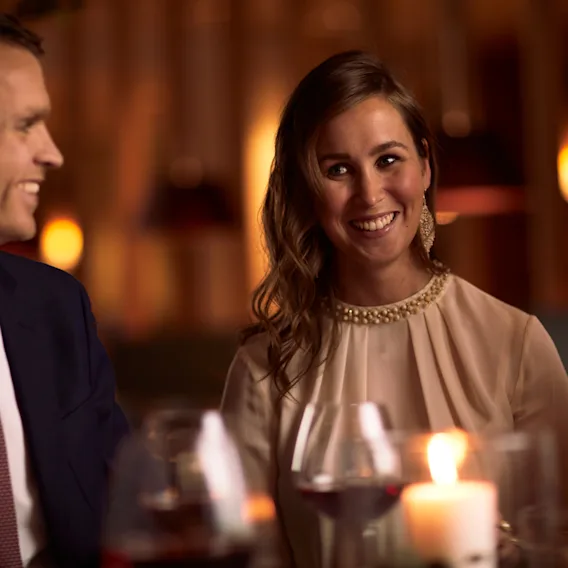 En mann og kvinne i pyntede klær smiler og ler sammen rundt et levende lys under en restaurantmiddag.