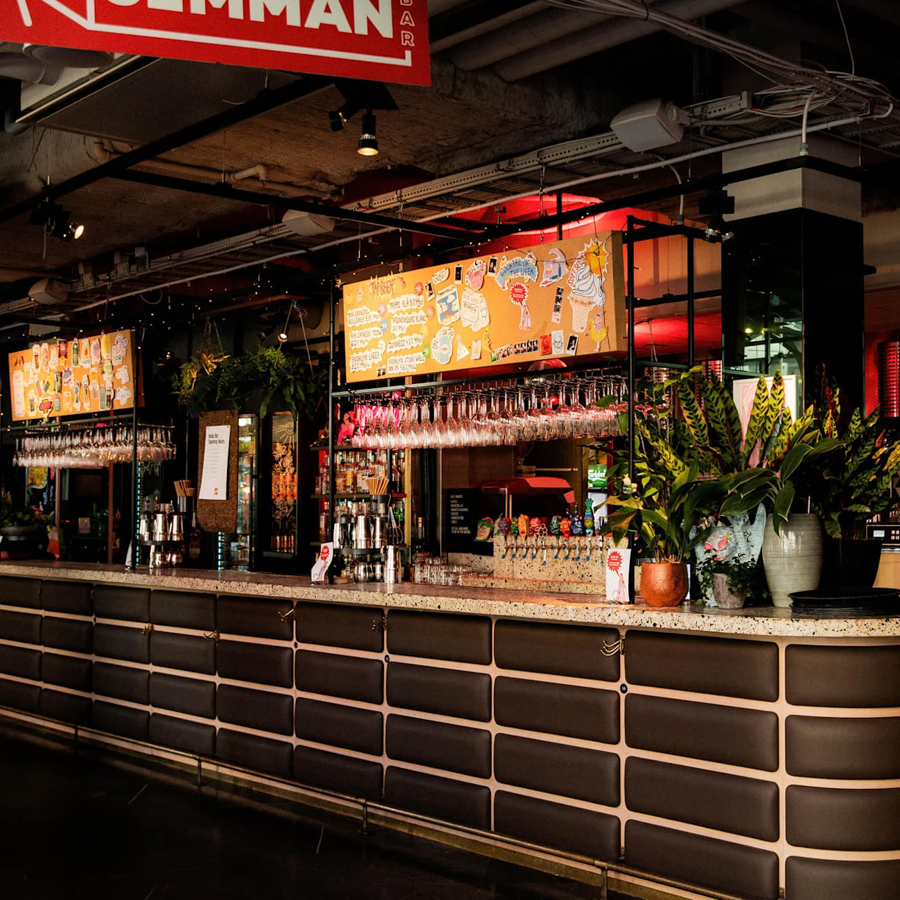 A bar with a curved counter displaying hanging wine glasses and potted plants. Bright orange signage with colorful illustrations hangs above. The setting is a modern, industrial-style space.