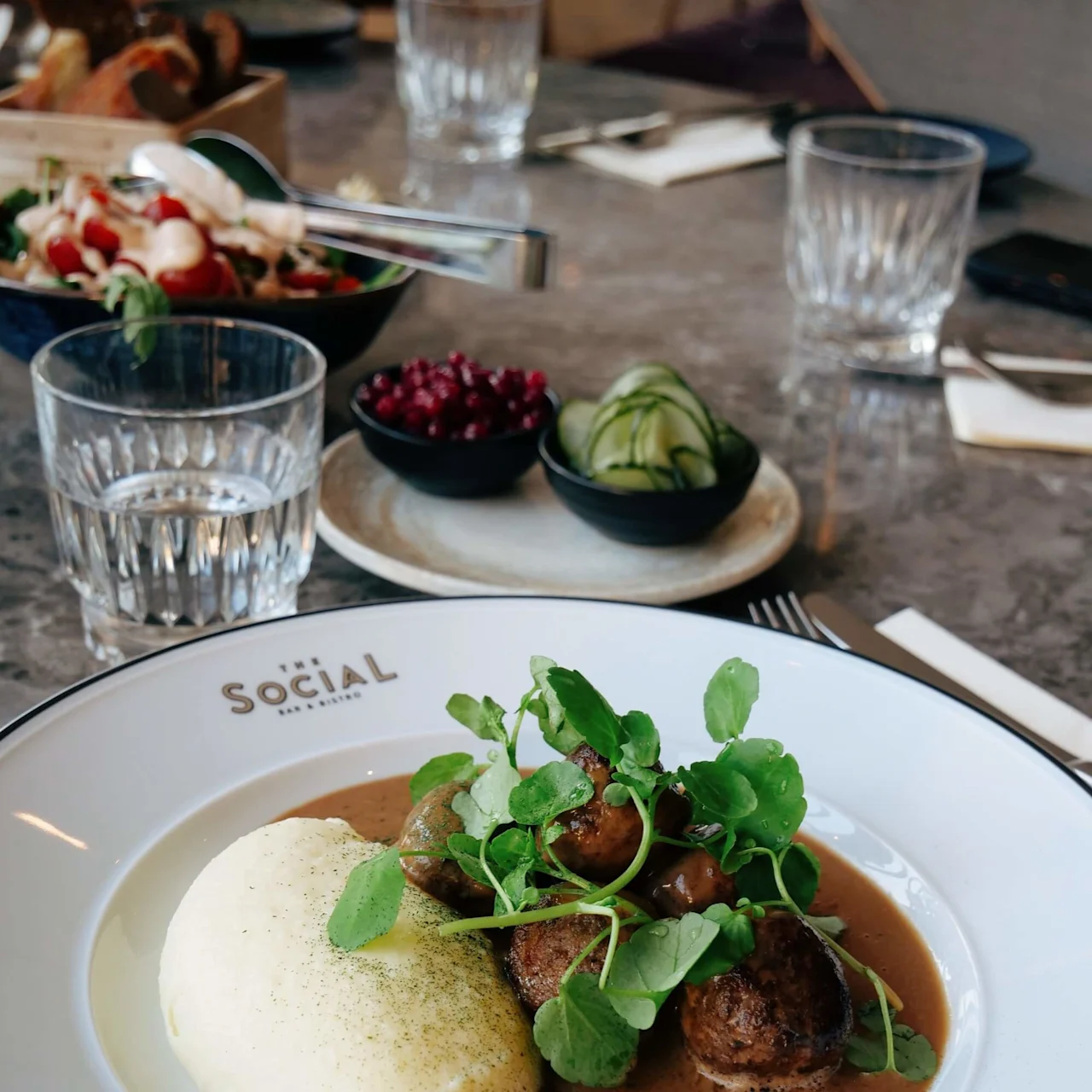 En tallrik med köttbullar i sås och potatismos, garnerad med grönt, står på ett restaurangbord. Nära finns rätter med lingon och pickles. Text: "The SOCIAL BAR & BISTRO."