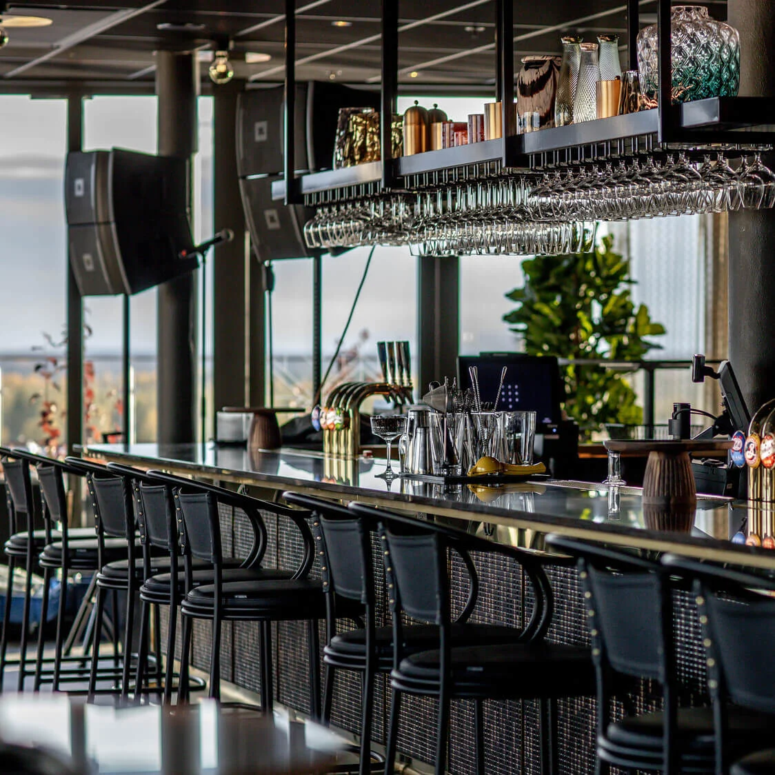 En moderne bar har en elegant bardisk med sorte stole, glas og spiritusflasker, placeret i et stilfuldt rum med store vinduer og dekorative hylder ovenover.