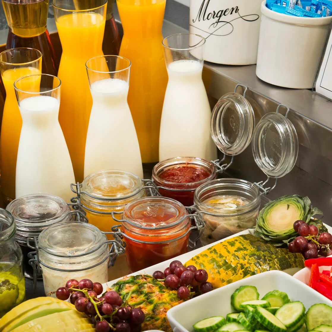 Ett urval av färska frukter, mjölk i genomskinliga karaffer och tillbehör i burkar visas upp för en bufféfrukost i en välbelyst miljö.