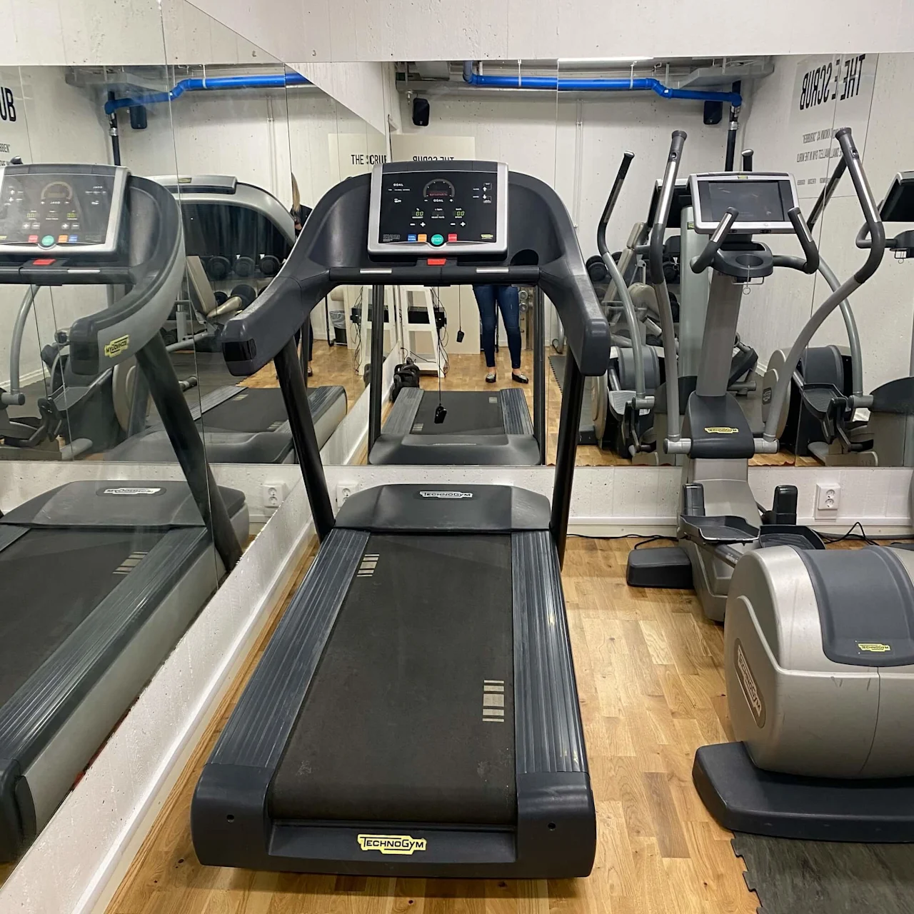 Et løbebånd med "TECHNOGYM"-mærkning står i et træningsmiljø, reflekteret i spejle på to sider med tilstødende konditionsudstyr og trægulv. Skilte siger "THE SCRUB HUB" og "SQUAD 208".