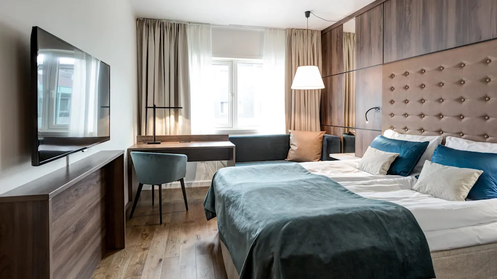 En pent redt seng med teal og hvitt sengetøy i et moderne hotellrom med veggmontert TV og skrivebord, opplyst av naturlig lys fra vinduet.