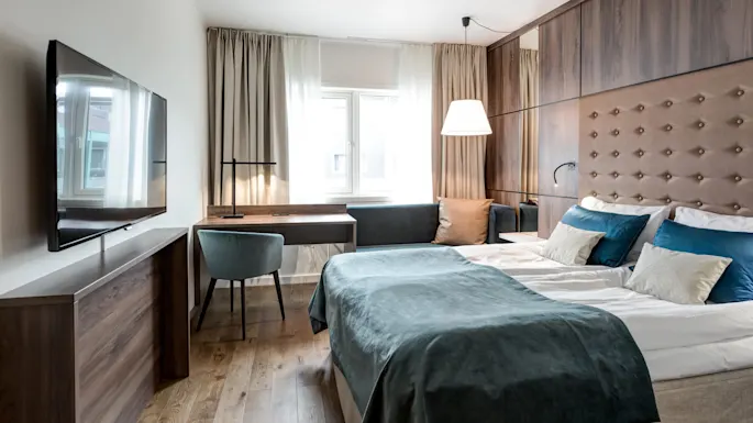 A neatly made bed with teal and white linens rests in a modern hotel room, featuring a wall-mounted TV and a desk, illuminated by natural light from the window.