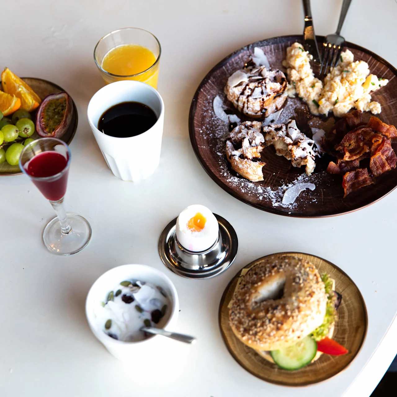 A breakfast spread includes a bagel with cucumber and bell pepper, scrambled eggs, bacon, pastries, coffee, orange juice, red beverage, fruit slices, and yogurt with seeds on a white table.
