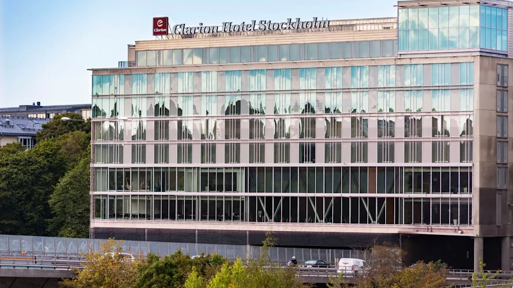 En modern hotellbyggnad visar skylten "Clarion Hotel Stockholm" ovanför reflekterande glasfönster. Träd och en väg med en bil finns i förgrunden.