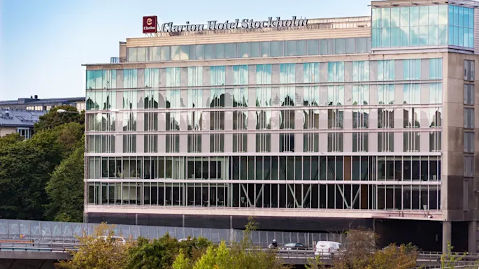 A modern hotel building displays the sign "Clarion Hotel Stockholm" atop reflective glass windows. Trees and a road with a car appear in the foreground.
