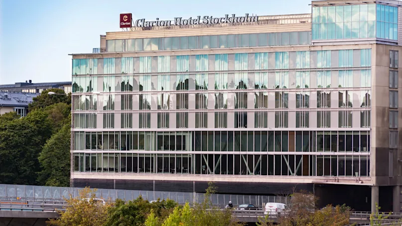 A modern hotel building displays the sign "Clarion Hotel Stockholm" atop reflective glass windows. Trees and a road with a car appear in the foreground.