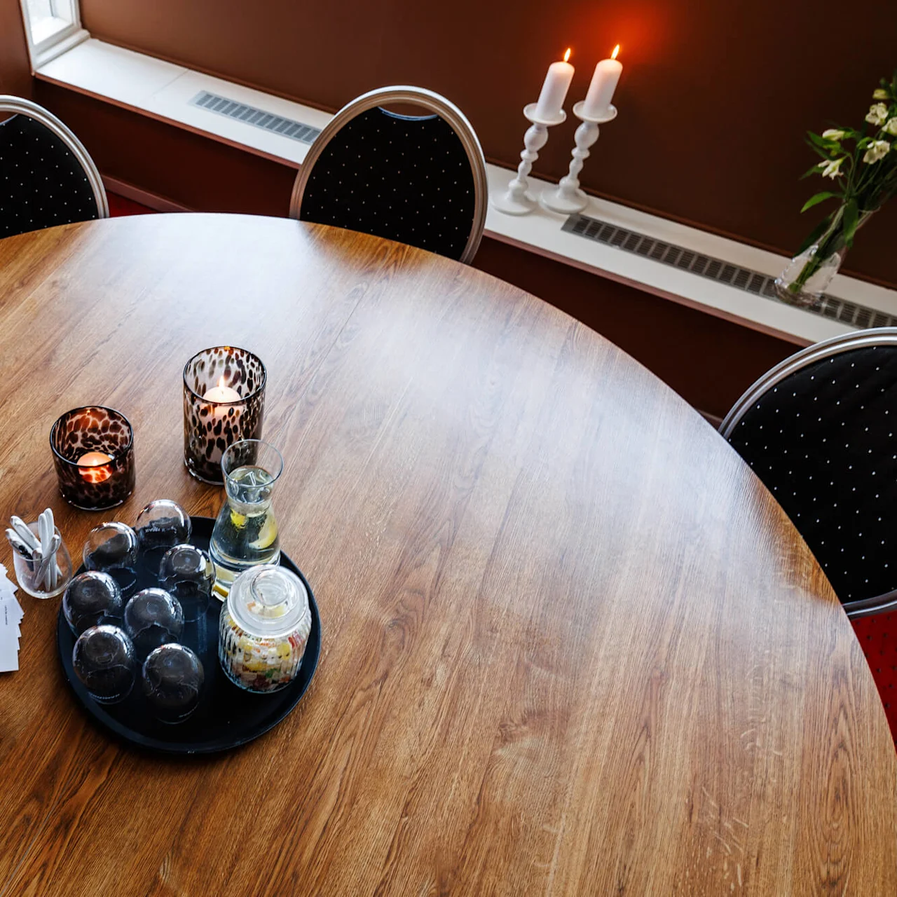 Runt träbord håller tända ljus, drycker och små desserter; kompletterat med servetter och en vas med vita blommor bredvid två vadderade stolar i ett mysigt rum.