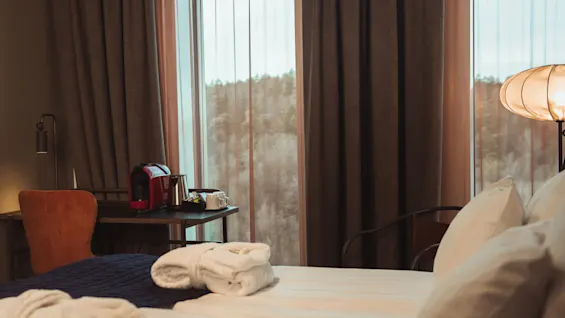 En säng med hopvikta morgonrockar ligger i ett mysigt hotellrum. Ett närliggande skrivbord har en röd kaffemaskin och vattenkokare, medan stora fönster visar en skogsvy.