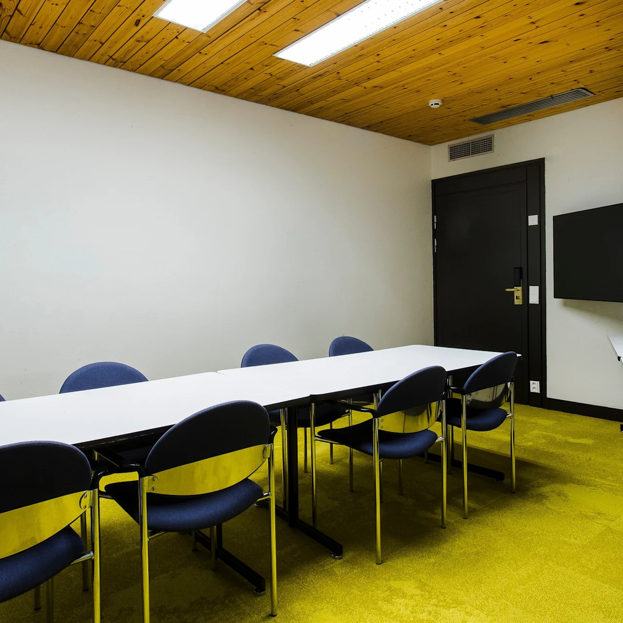 A long table with eight blue chairs sits in a small conference room with a wooden ceiling. A TV is mounted on the wall, and a side table holds a water pitcher and glasses.