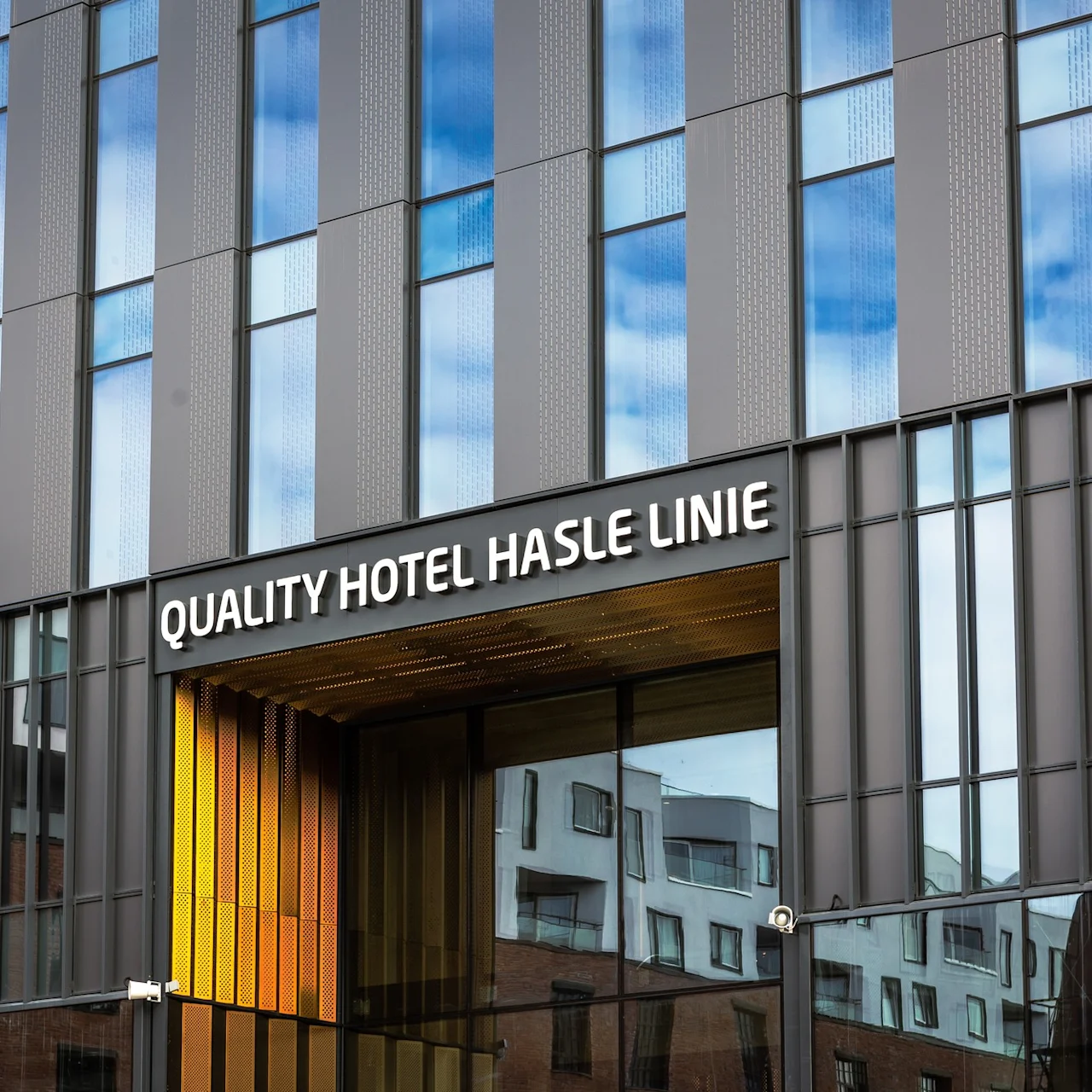 En modern hotellentré har stora glasfönster som speglar blå himmel och intilliggande byggnader. Skylten lyder ”QUALITY HOTEL HASLE LINIE” ovanför ingången med en gul accent.