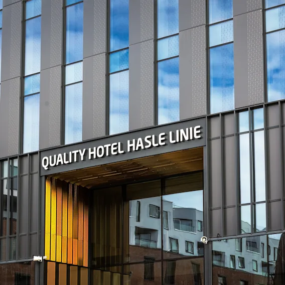 En moderne hotellinngang har store glassvinduer som gjenspeiler blå himmel og nærliggende bygninger. Skiltet lyder “QUALITY HOTEL HASLE LINIE” over inngangen med en gul aksent.