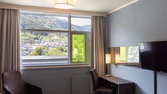 Ett hotellrum har två stolar och ett bord bredvid ett skrivbord med en lampa. Ett stort fönster erbjuder en naturskön utsikt över bergen och en stad nedanför.