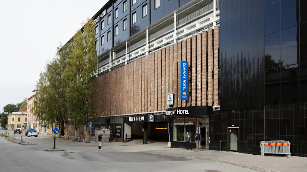 Comfort Hotel building with a brick facade and blue sign. People walk nearby on the street, with trees and a parking entrance. Visible text: "Comfort Hotel", "völkommen till vår webshöp".