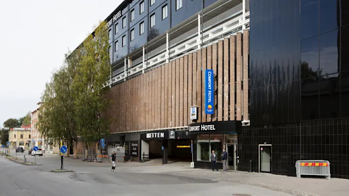 Comfort Hotel-bygning med murfasade og blått skilt. Folk går i nærheten på gaten, med trær og parkeringsinngang. Synlig tekst: "Comfort Hotel", "völkommen till vår webshöp".