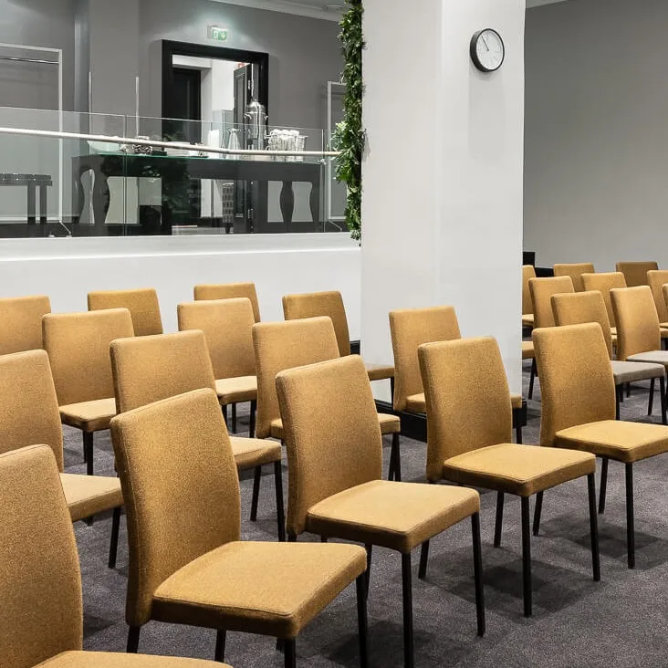 Rader av beige stoler står pent arrangert i et konferanserom med grå vegger, en klokke og projektorer i taket. En hevet plattform med glassrekkverk har forfriskninger. Nødutgangsskilt er synlige.