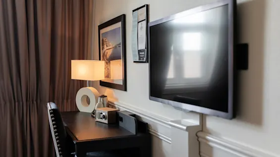 En komfortabel hotelværelsesopsætning med et skrivebord, lampe, vægmonteret TV og en informationsskærm.