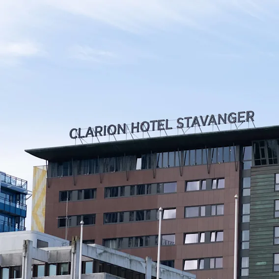 Clarion Hotel Stavanger-skiltet sitter på toppen av en moderne, fleretasjes bygning med brune og grønne paneler, omgitt av en klar blå himmel.