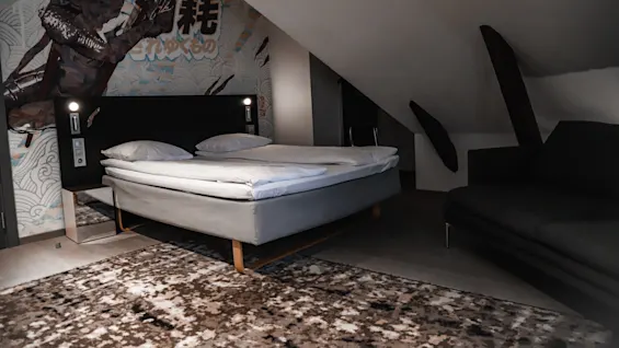 A bed with white linens is placed under a slanted ceiling, next to a dark sofa; walls feature artistic artwork, including Japanese characters.