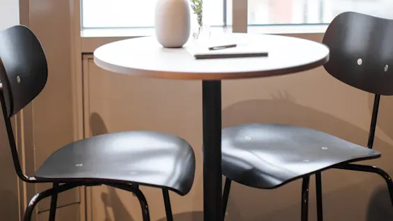 A small round table with a white vase and plant sits between two black chairs, in a room with a large window showing a blurred cityscape outside.