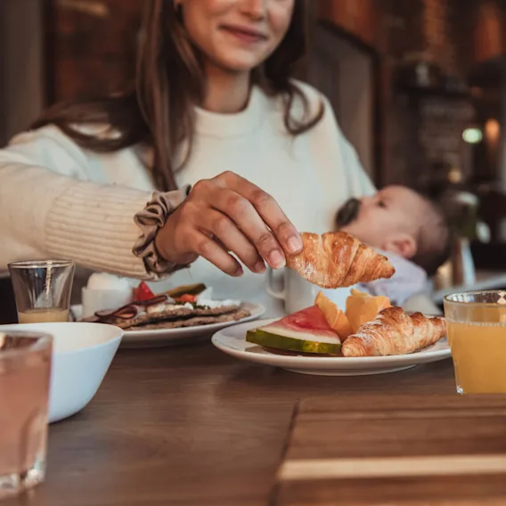 Kvinna håller en croissant, sitter vid ett bord med en bebis i famnen. Olika frukostartiklar och drycker på träbordet, i en mysig kafémiljö.