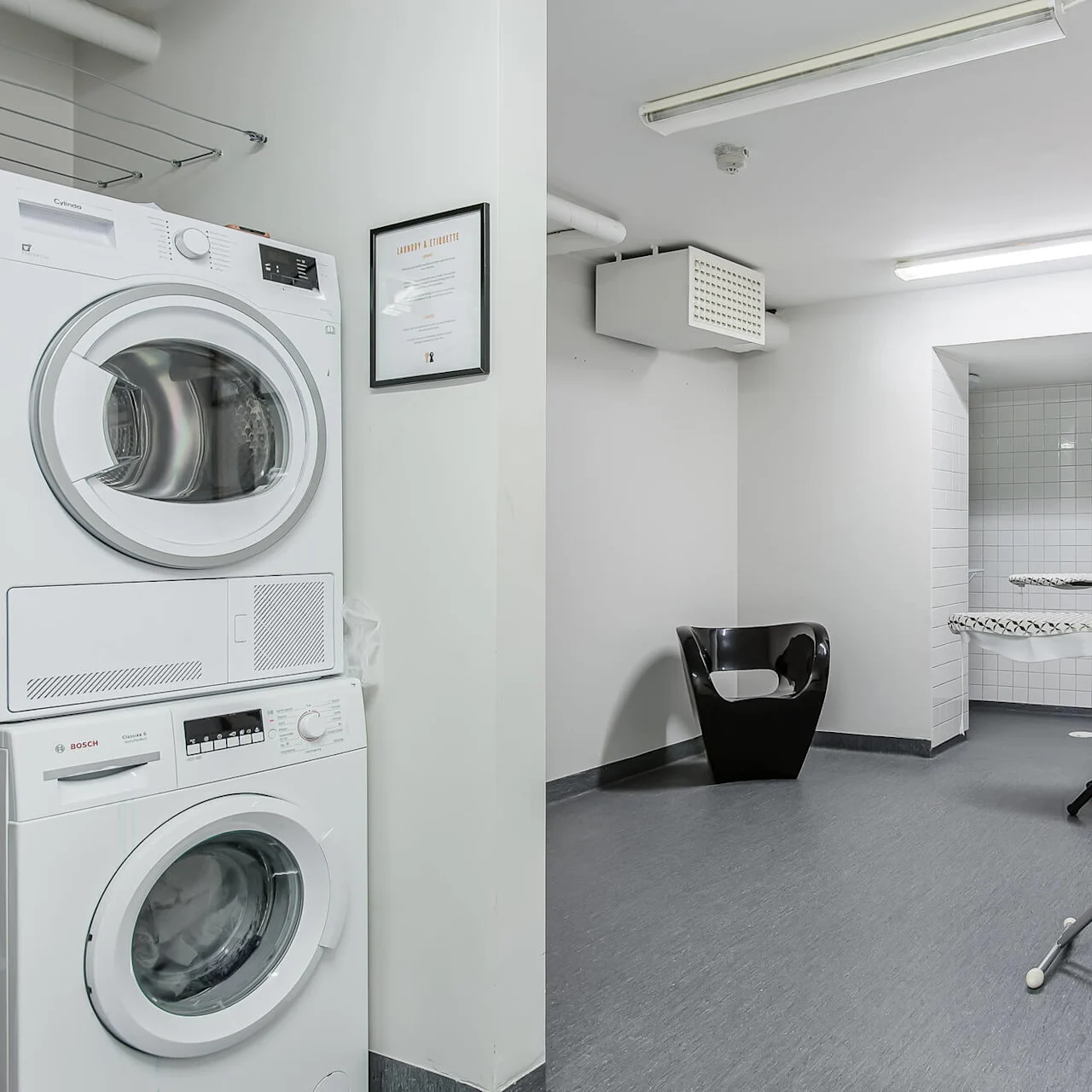 Pesu- ja kuivaimet pinottu silittämisalueen viereen, jossa on kaksi silityslautaa ja -rautaa. Moderni tuoli on lähellä kirkkaan loisteputkivalon alla. Seinällä on kehystetty "Laundry Ettiquette" -ilmoitus.