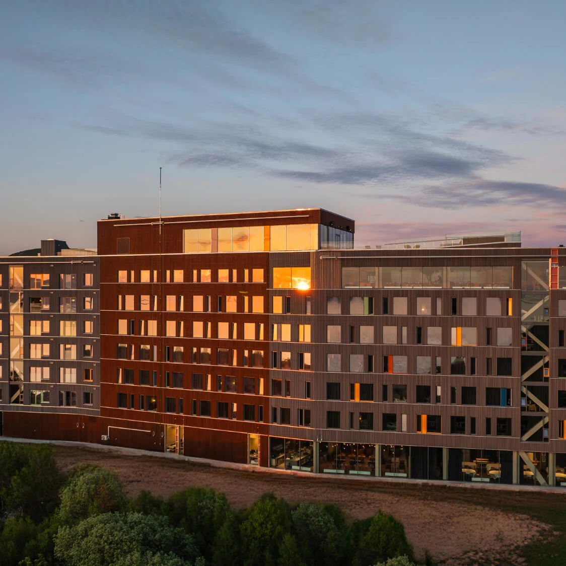 A modern multi-story wooden building reflects the sunset, with mountains and a fjord in the background. It stands surrounded by lush greenery.