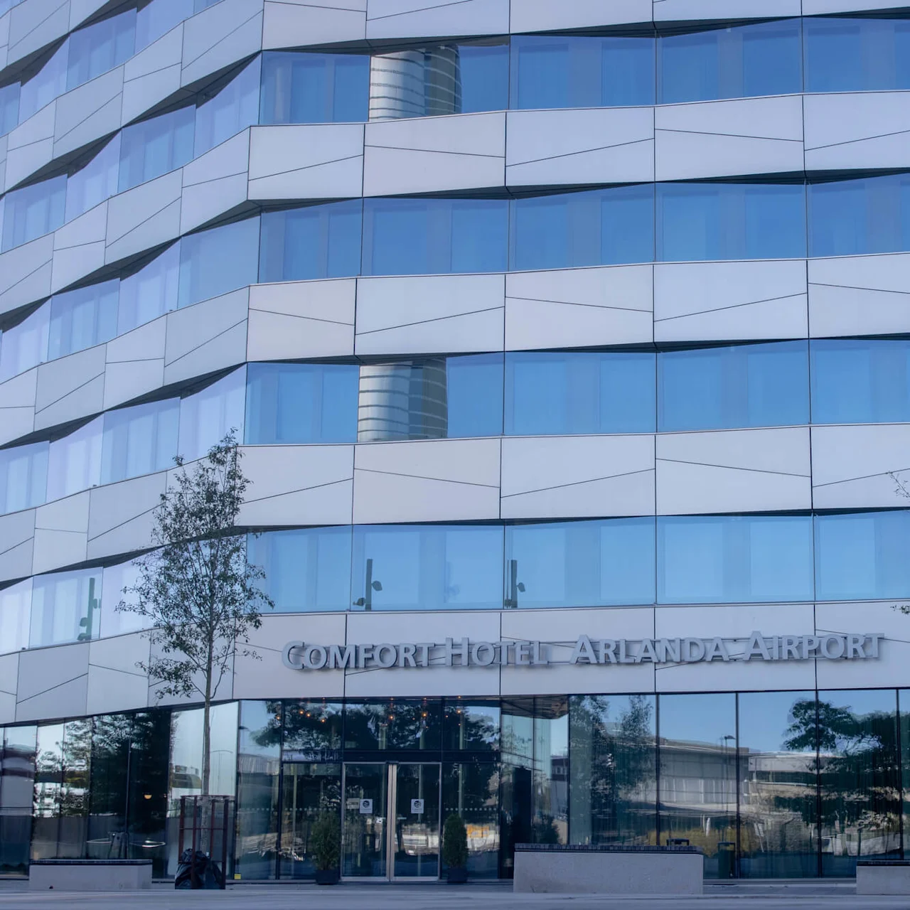 A modern glass building facade reflects the sky. Trees and a symmetrical design border the entrance. Text reads: "COMFORT HOTEL ARLANDA AIRPORT."
