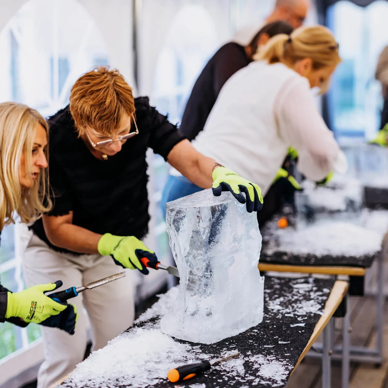 People carve ice blocks using chisels on tables in a tent, wearing gloves, with onlookers in the background.