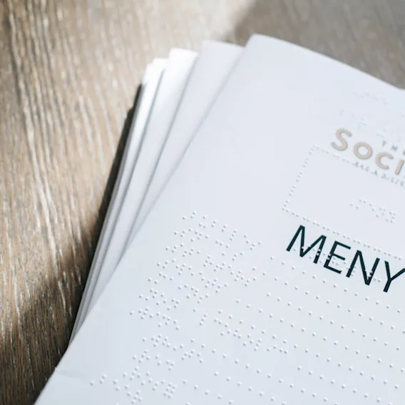 En papir-menymed punktskrift ligger på en treoverflate. Teksten på menyen lyder "THE SOCIAL ALL IN. OUTED. MENY".