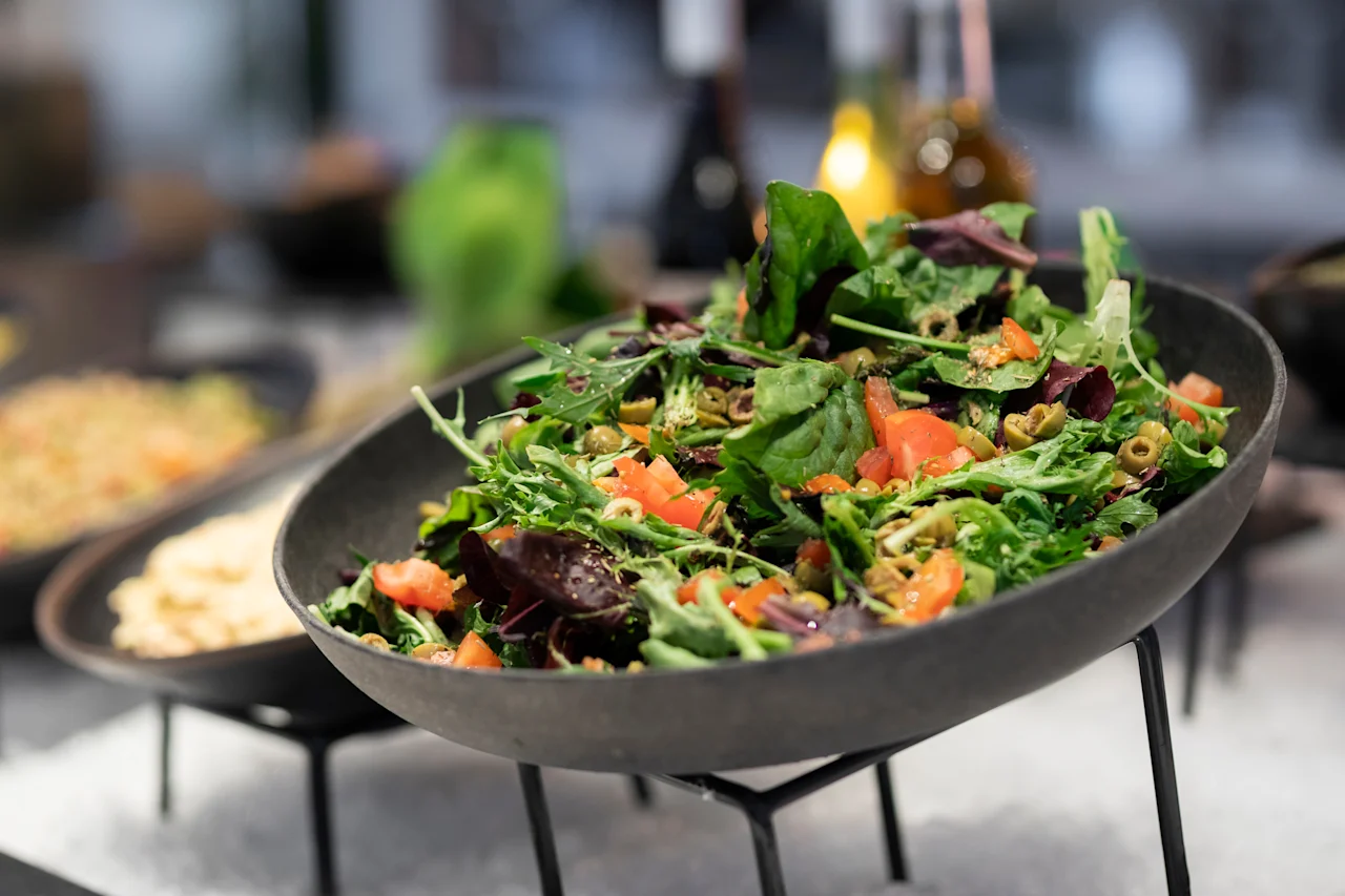 Tuore salaatti, jossa on sekoitettuja vihreitä, tomaatteja ja oliiveja tummassa kulhossa noutopöydässä.