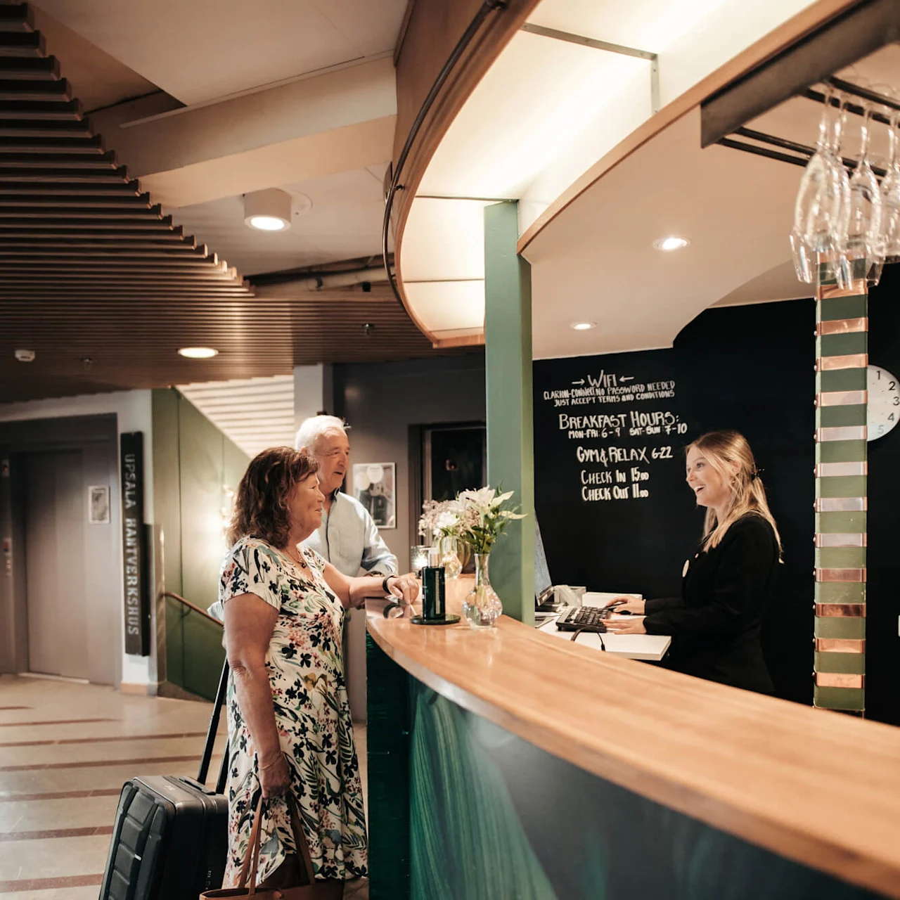 Two guests are checking in at a hotel reception desk, greeted by a staff member in a well-lit lobby.