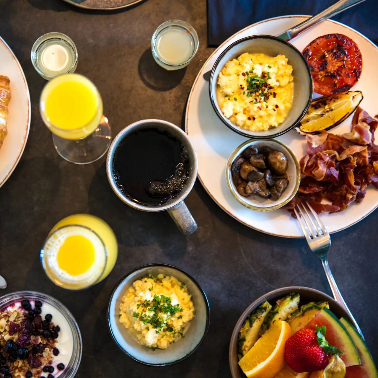 Various breakfast dishes arrayed on a dark table, featuring eggs, fruit, pastries, and beverages.