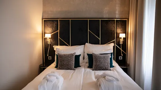 En luksuriøs hotelseng med sprøde hvide lagner, bløde puder og to badekåber, oplyst af varme sengelamper.