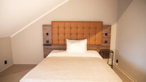 Säng med beige sängkläder och en vit kudde står mot ett brunt vadderat sänggavel. Två väggmonterade lampor flankerar sänggaveln i ett mysigt rum med sluttande tak.