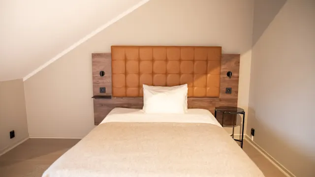 Seng med beige sengetøy og en hvit pute står mot en brun polstret sengegavl. To veggmonterte lamper flankerer sengegavlen i et koselig rom med skrånende tak.