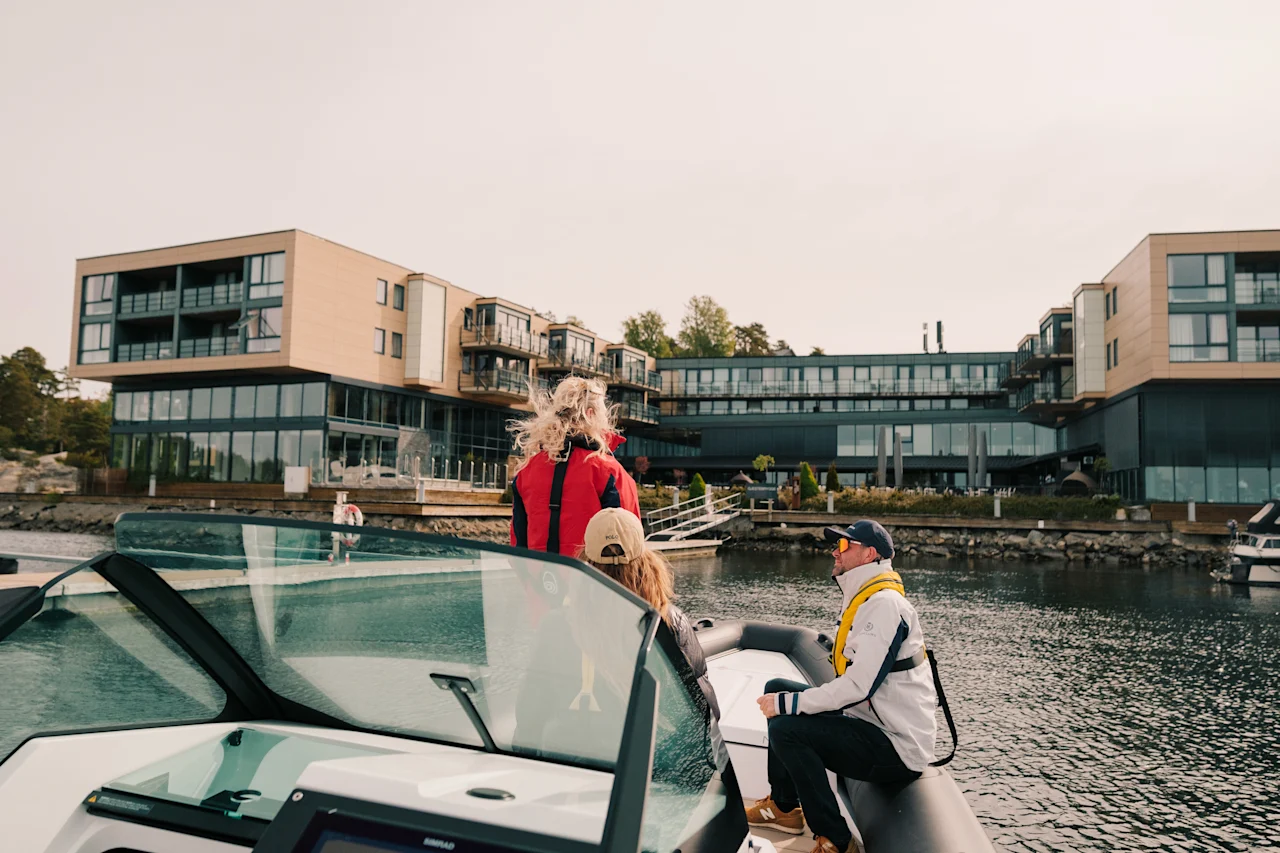 Bådfolk nyder en naturskøn tur mod et moderne hotel ved vandet, med en klar himmel over.