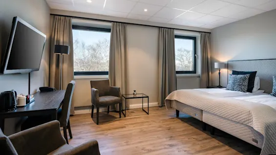 Et pænt arrangeret hotelværelse med en seng med mønstrede puder og en dyne, et vægmonteret TV og et skrivebord med en kedel. Store vinduer giver udsigt til træer udenfor.