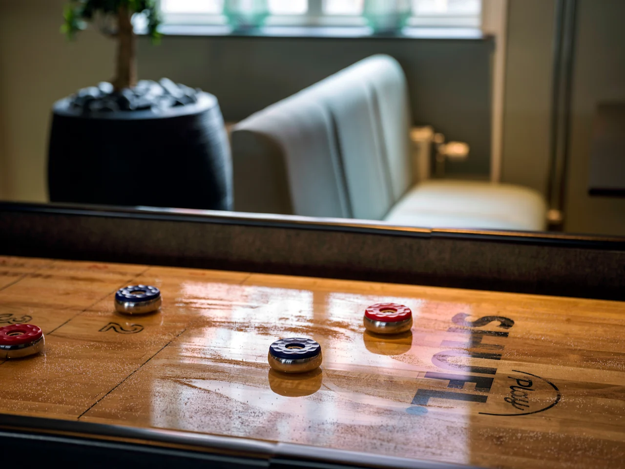 A wooden shuffleboard table with red and blue pucks, ready for a game. The words SHUFFLE and Play are visible on the table.