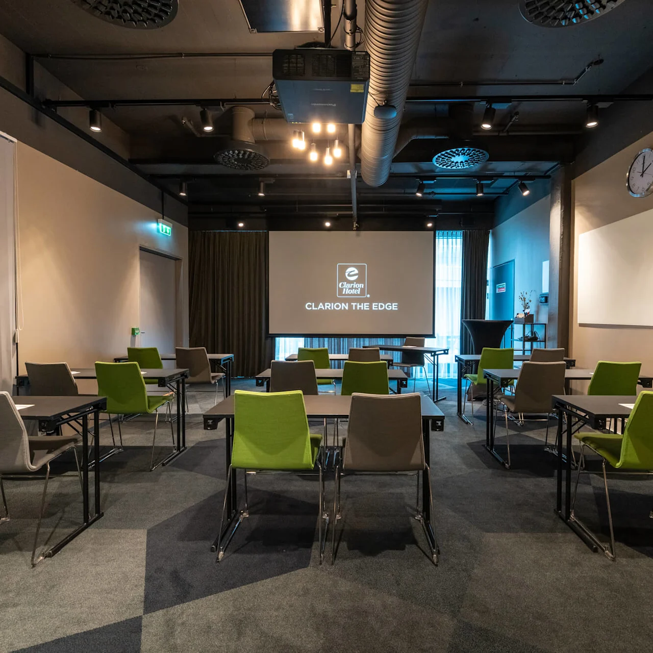 Projektor viser "Clarion The Edge" i et konferencelokale med grønne og grå stole, borde, loftsbelysning, gardinbaggrund og en vægur.