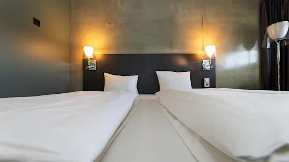 A neatly made double bed with white linens. Two pillows rest on a dark headboard under wall-mounted lamps, creating a cozy ambiance in a modern bedroom.