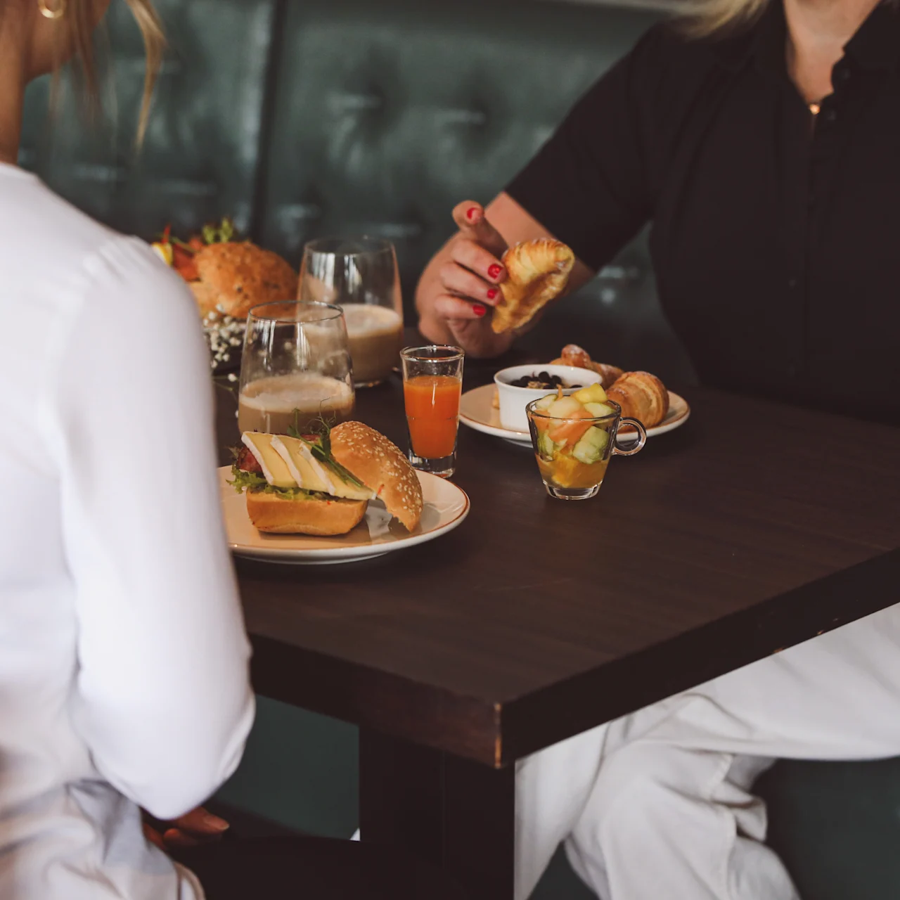 Two people sit at a wooden table enjoying lunch. Sandwiches, coffee, juice, and fruit are served. They are in a casual dining setting with green upholstered seating.