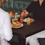 Two people sit at a wooden table enjoying lunch. Sandwiches, coffee, juice, and fruit are served. They are in a casual dining setting with green upholstered seating.