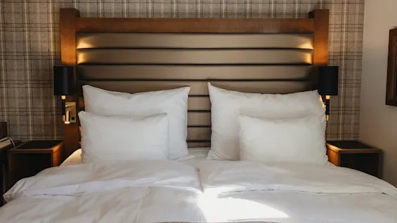 Et hyggeligt hotelværelse med en pænt redt seng, hvidt sengetøj og en stilfuld sengegavl. Sollys strømmer ind over sengen.